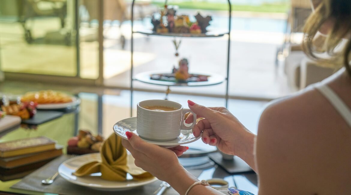 Mulher segurando uma xicara com café com leite vegano em sua frente uma mesa posta com variedade de pães, salgados, boles e doces veganos para tomar seu café da manha em um hotel maravilhoso