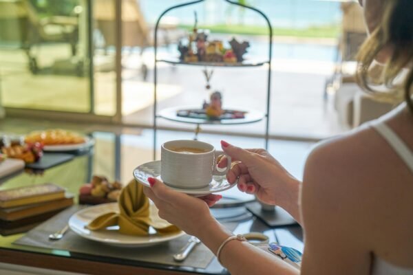 Mulher segurando uma xicara com café com leite vegano em sua frente uma mesa posta com variedade de pães, salgados, boles e doces veganos para tomar seu café da manha em um hotel maravilhoso