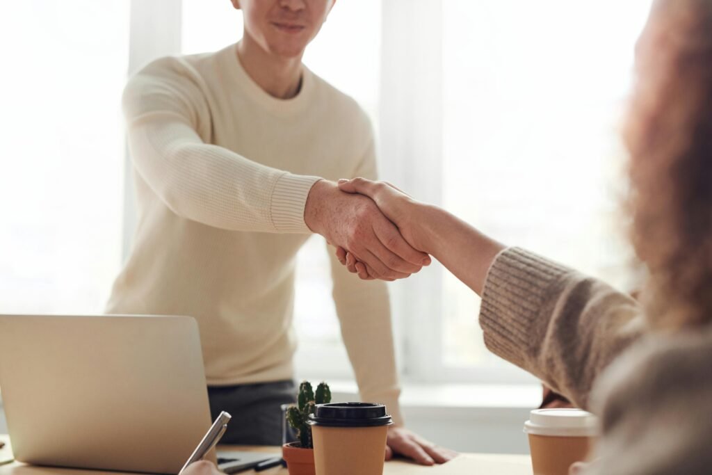 Homem em pé em frente a mesa, com computador e um café apertando a mão de uma mulher, sentada à mesa com um copo de café a sua frente, ambos fecharam um acordo.