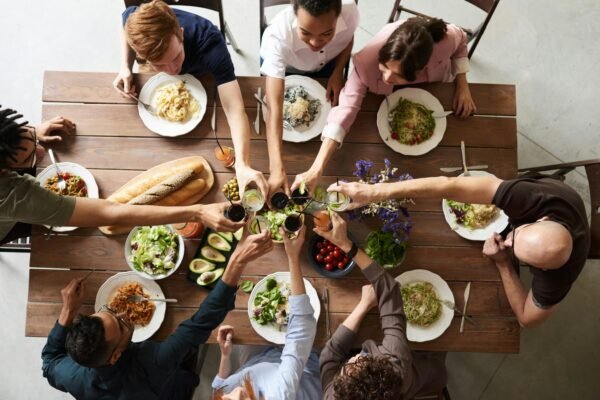 Mesa posta com os pratos servidos, cada prato contém alimentos diferentes, mostrando a diversidade alimentar. As pessoas estão sentadas à mesa em frente aos seus respectivos pratos brindado a oportunidade de estarem juntas, indiferente do que cada um se alimenta.